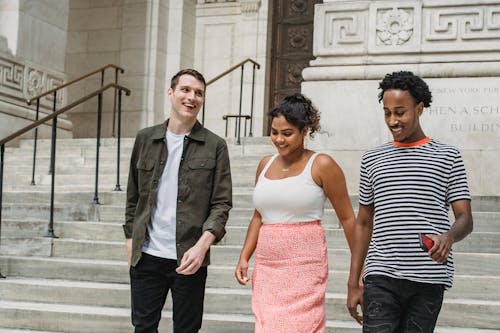 Group of young positive multiracial friends in summer clothes strolling on stairs with phone in hand in city street near building in daytime