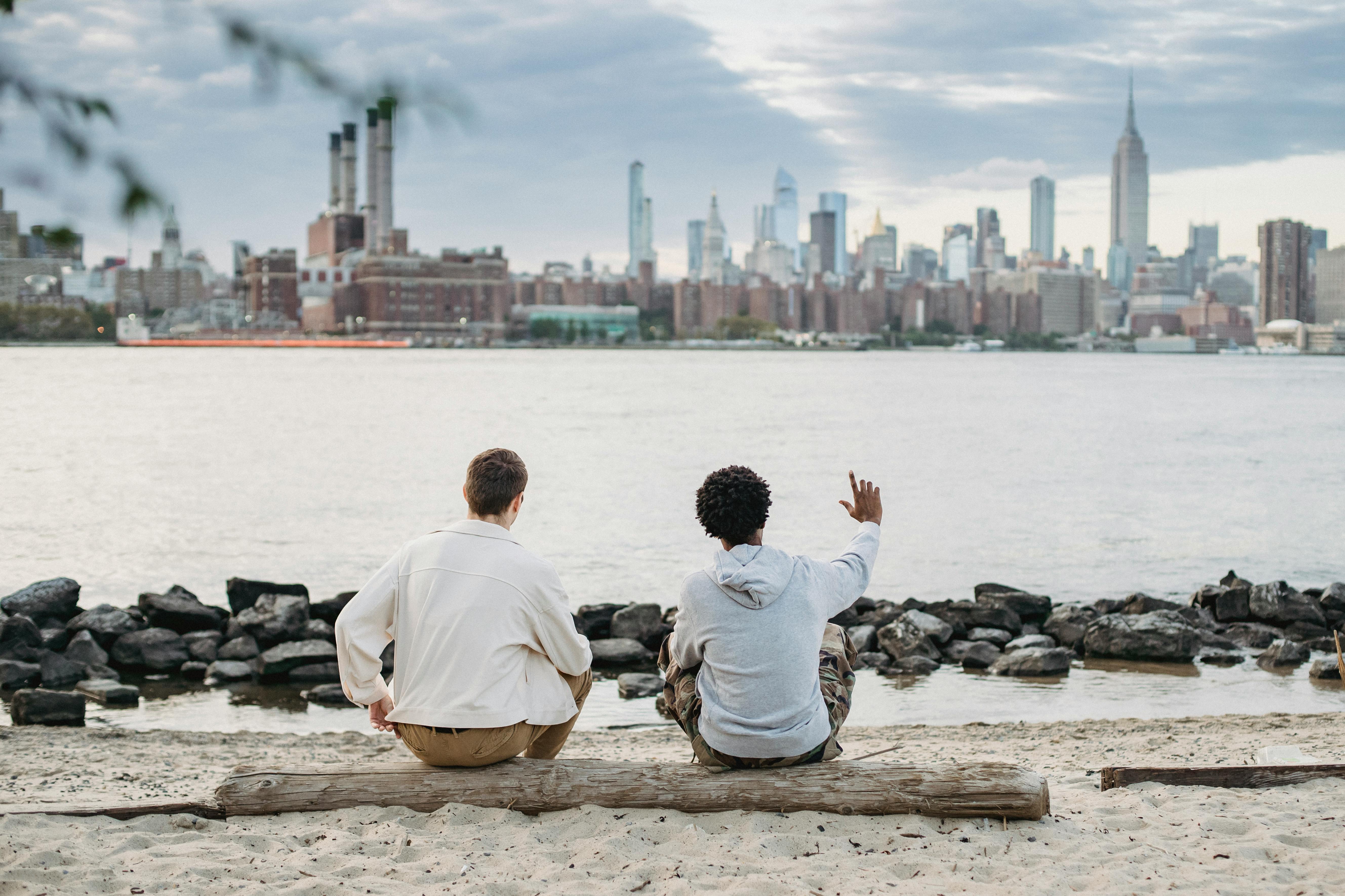 unrecognizable diverse friends sitting on bank of river against cityscape