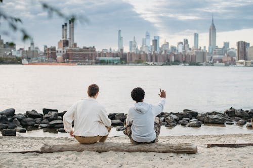 Free Back view of crop anonymous multiracial friends sitting together on wooden trunk on bank of river and looking at urban landscape Stock Photo