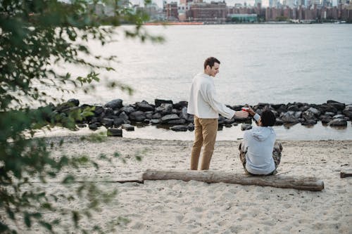 Positive young guy giving drink in takeaway cup to faceless black male friend sitting on wooden trunk on seashore with urban landscape in daytime