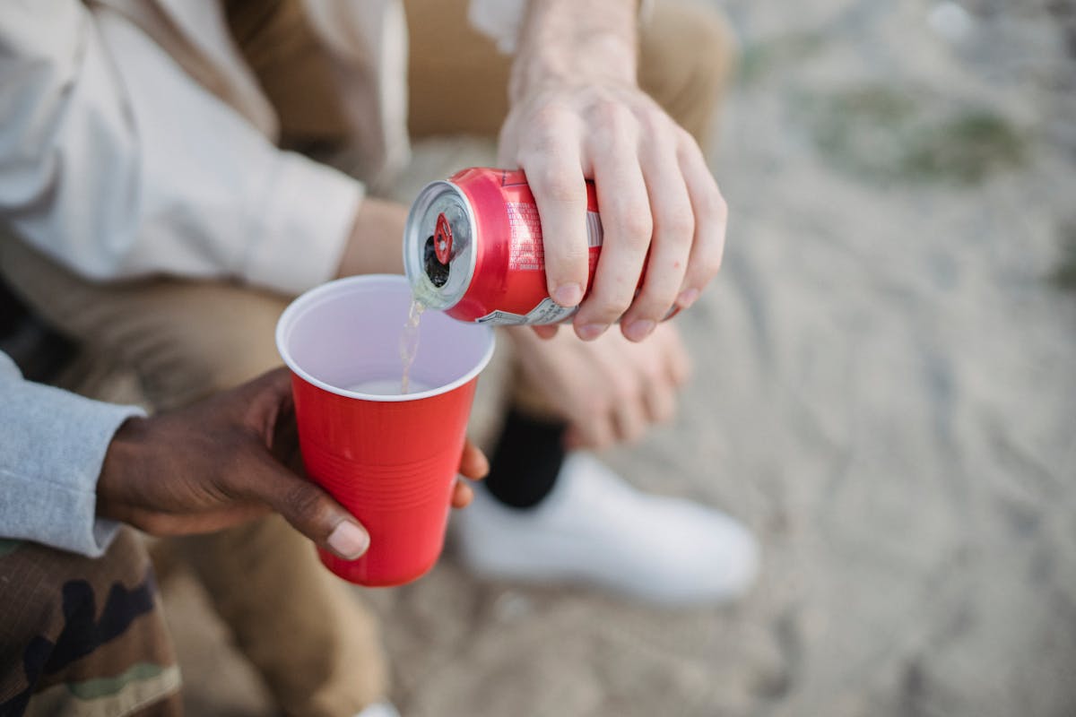 From above of crop anonymous man pouring fizzy drink from can into red plastic cup of black friend while sitting on sandy ground