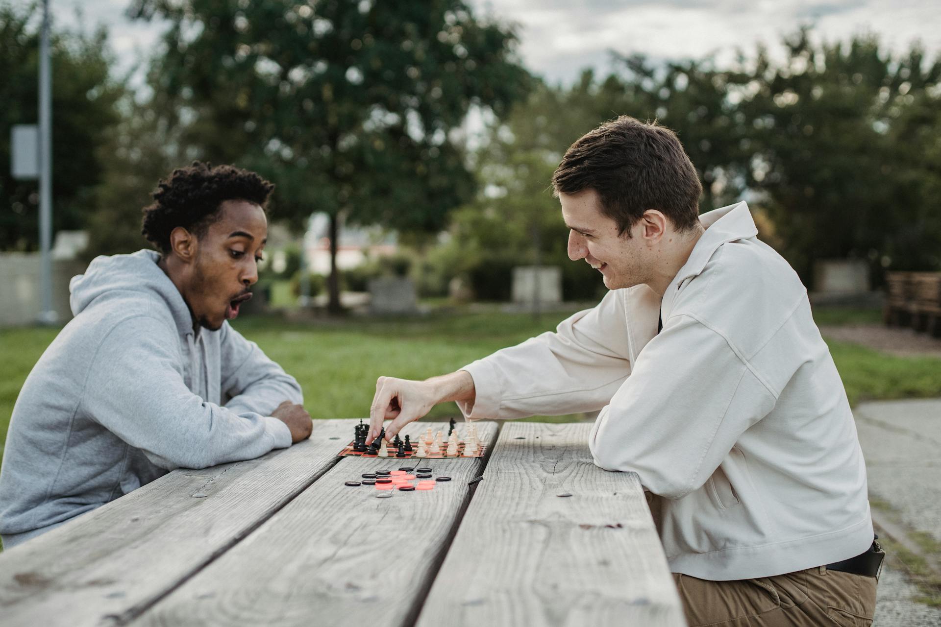 Side view of smiling young multiracial male friends sitting at wooden table in city park and enjoying interesting chess game