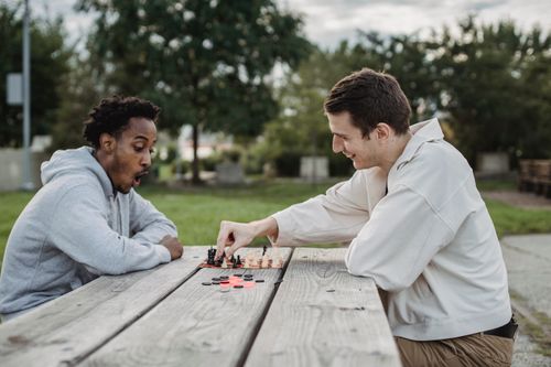 Free Side view of smiling young multiracial male friends sitting at wooden table in city park and enjoying interesting chess game Stock Photo