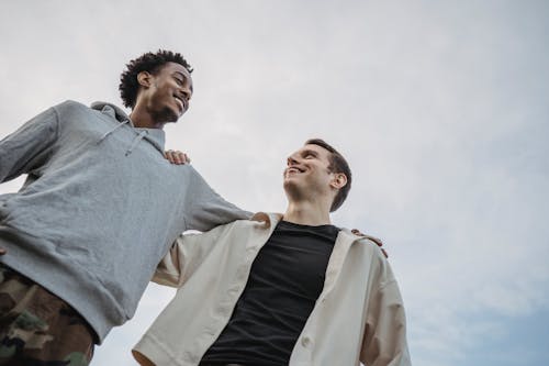Happy young multiethnic male friends side hugging against cloudy sky