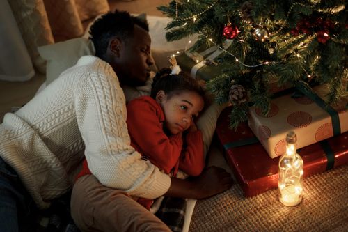 Free Father and daughter resting together under a Christmas tree with glowing lights and gifts. Stock Photo
