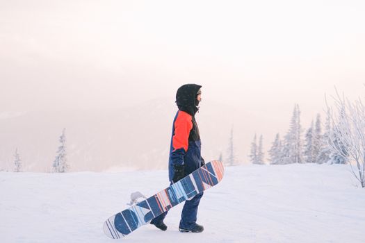 Teen snowboarder walking with a snowboard in a snowy mountain landscape under a bright sky.