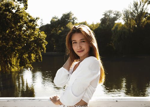Free A smiling young woman in a white outfit poses on a bridge by a serene lake, surrounded by lush greenery. Stock Photo