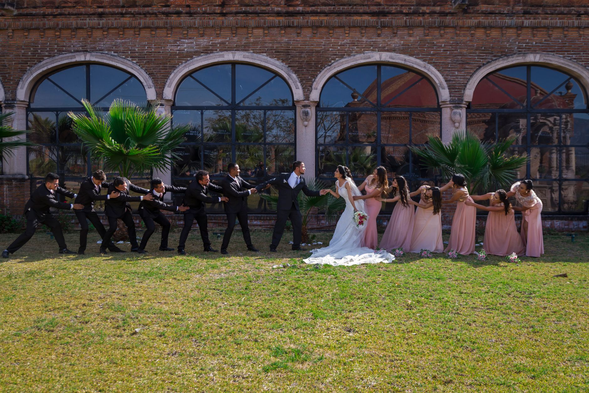 Bride and groom in wedding attire with bridal party playing tug of war outdoors.