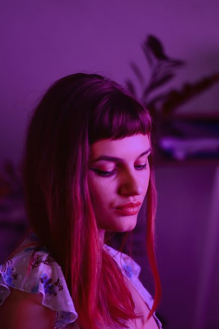 Thoughtful young female in casual clothes with long hair looking down in dark room with purple neon ligh