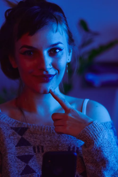Positive young female in warm woolen sweater in dark room illuminated with violet neon light touching chin and looking at camera