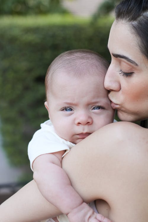Crop woman mother with dark hair kissing infant baby with bright blue eyes on street in daytime