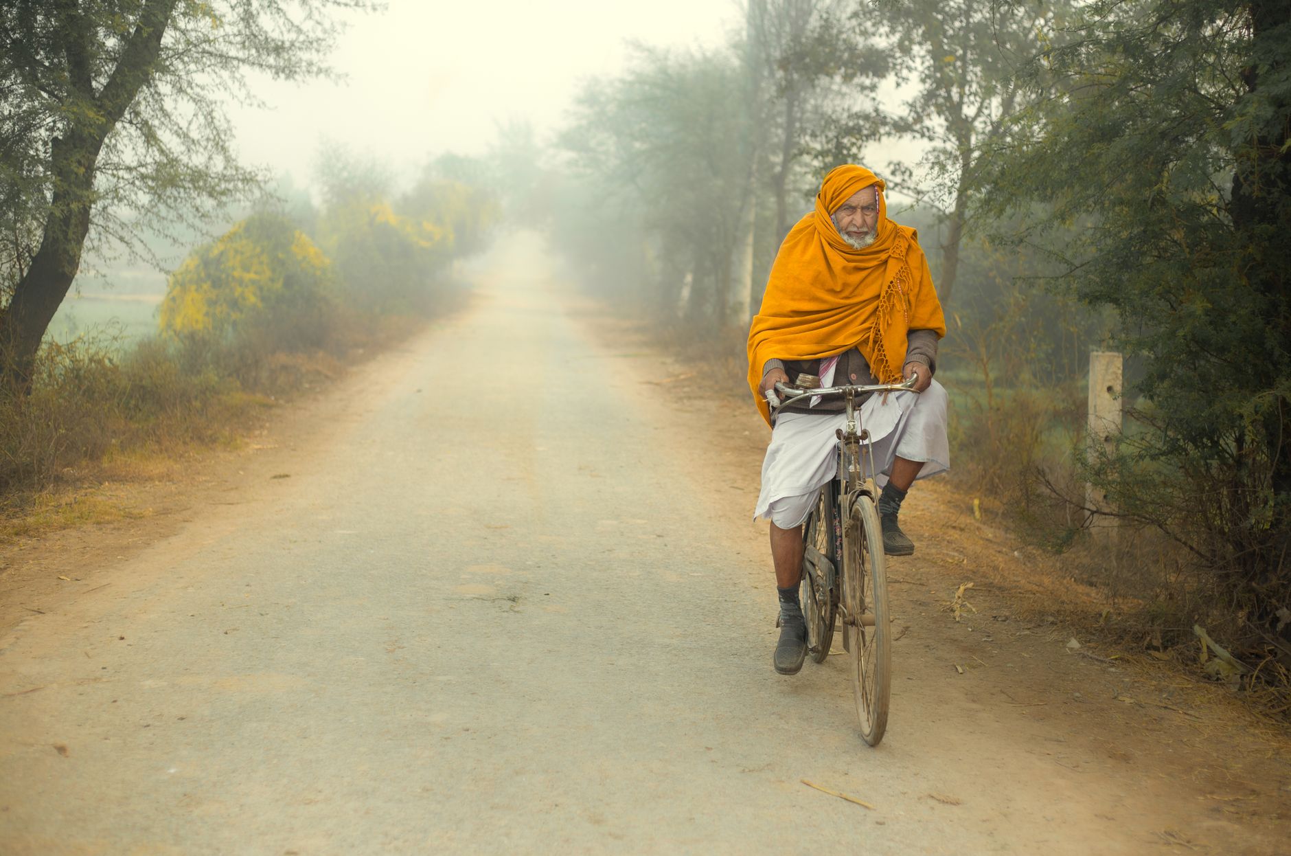 An elderly man in an orange headscarf rides a bicycle on a foggy rural road surrounded by trees.
