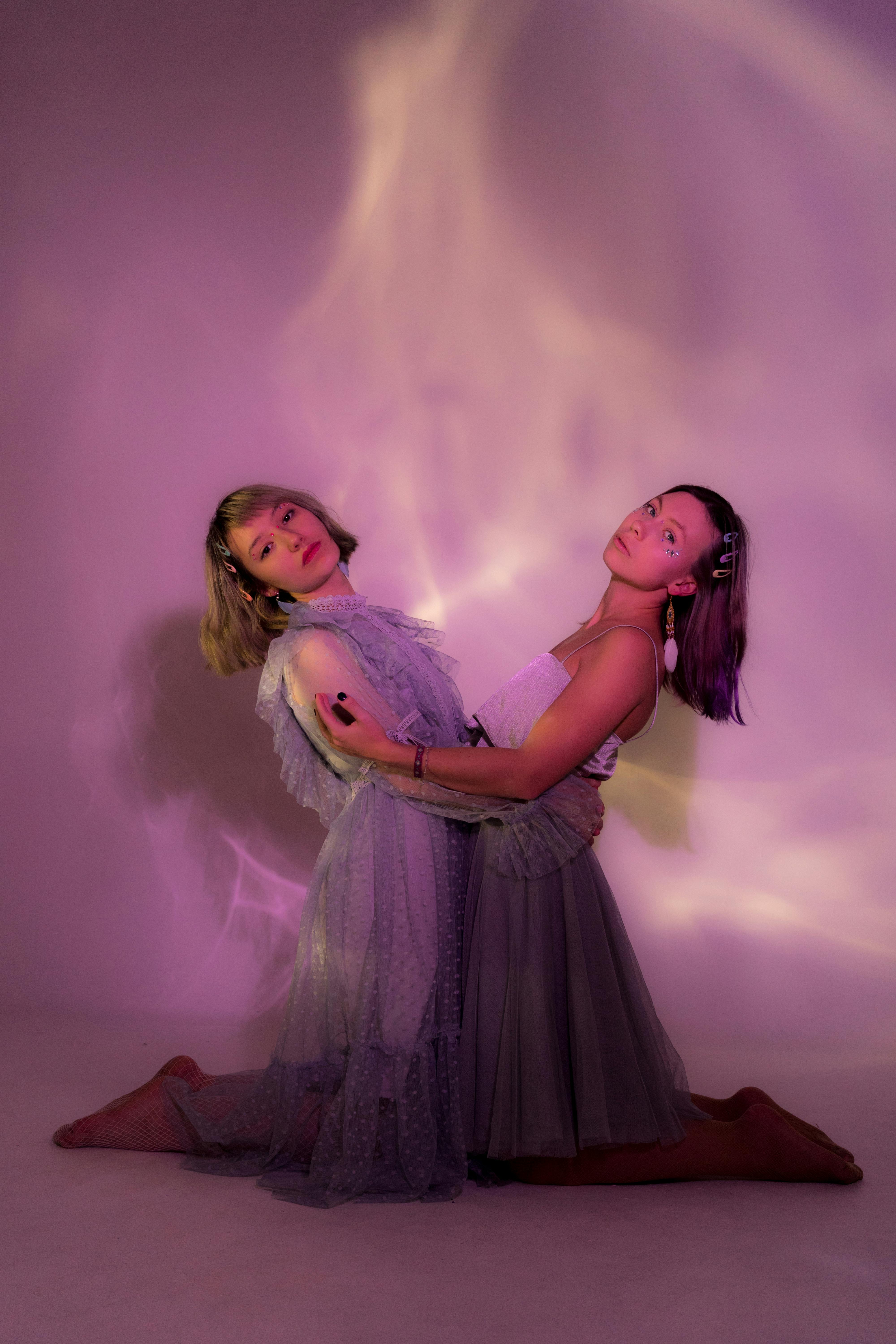 Full body of stylish female friends wearing elegant outfits looking at camera and embracing while standing on knees in studio