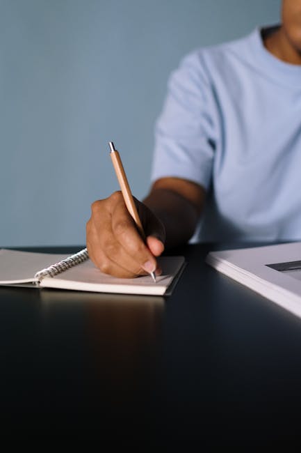 A person writing in a notebook with a pen on a dark surface, focusing on the hand.