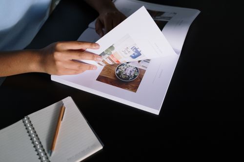 Free A person flipping through a glossy magazine on a desk with a notepad and pencil nearby. Stock Photo