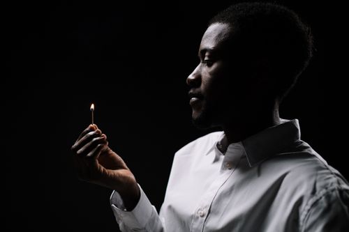 Free A contemplative man in white shirt holding a lit match against a dark background. Stock Photo