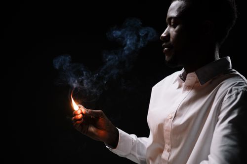 Free Side view of a man in a white shirt lighting a matchstick with smoke in a dark room. Stock Photo