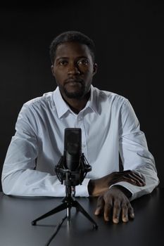 A serious black man in a white shirt sits at a table with a microphone, against a dark background.