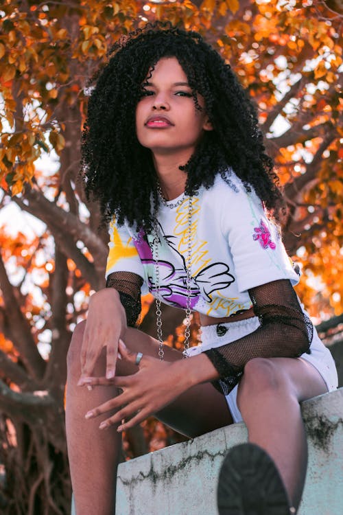 Free Low angle of confident young African American lady wearing colorful shirt sitting on shabby metal construction against autumn trees and looking at camera Stock Photo