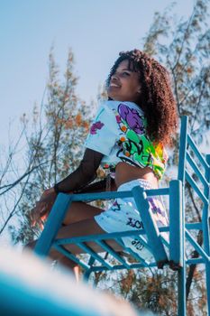 From below of smiling youngster black lady wearing informal cloth sitting in metal armchair in park and looking away on sunny autumn day