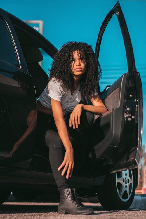 Low angle of stylish young African American lady wearing black trousers with boots leaning on door of black car and looking at camera