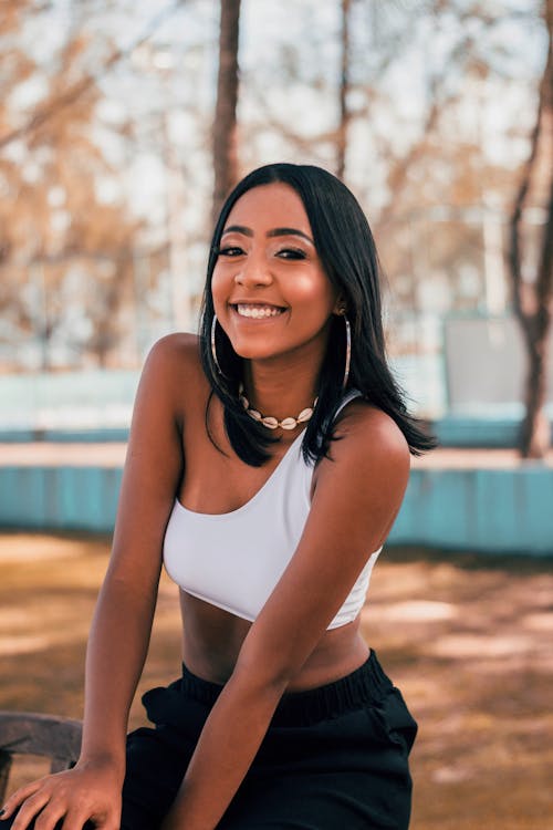 Free Delighted young African American lady with long black hair resting in urban park on sunny day and smiling at camera Stock Photo