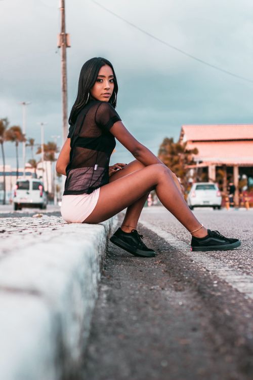 Free Side view of confident ethnic woman wearing transparent t shirt sitting on concrete barrier near road and looking at camera in daylight Stock Photo