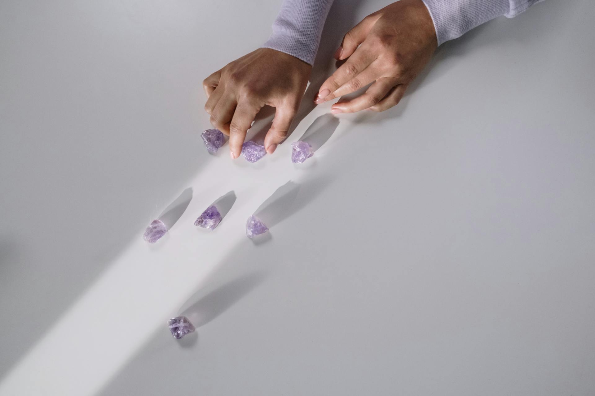 Hands arranging amethyst crystals on a white surface, creating patterns with light and shadows.