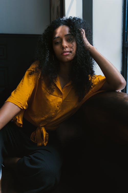 Young thoughtful African American woman in trendy wear and with curly hair relaxing sitting in armchair beside window and looking at camera