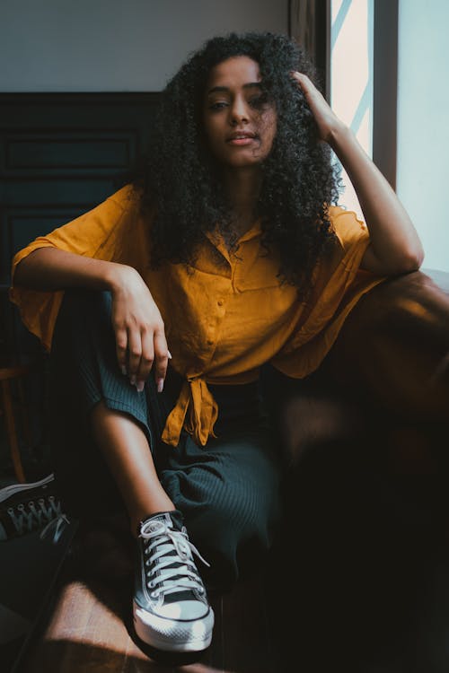 Young African American female in smart casual outfit and sneakers sitting in armchair with legs crossed and looking at camera