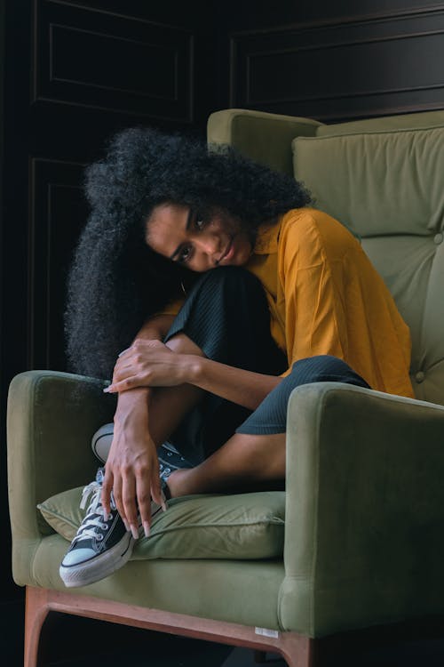 Free Young African American female with curly hair and casual outfit sitting in cosy armchair and looking at camera in apartment Stock Photo