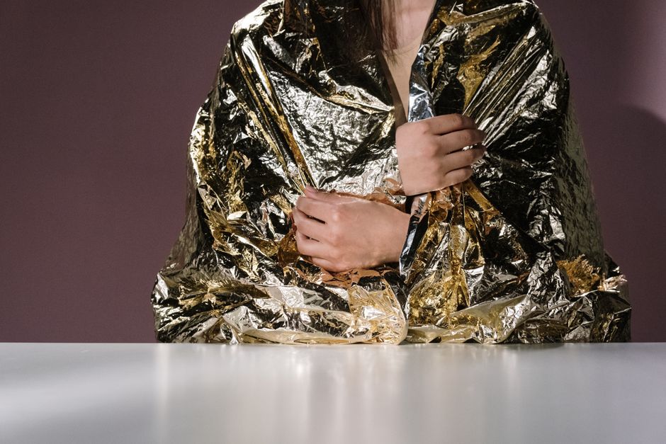 A person wrapped in golden metallic material sitting at a table, showcasing fashion and conceptual art.