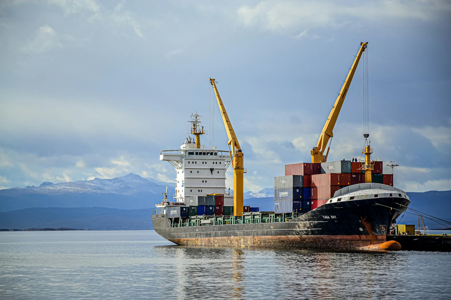 A large cargo ship at port with tower cranes and stacked containers against a mountain background.