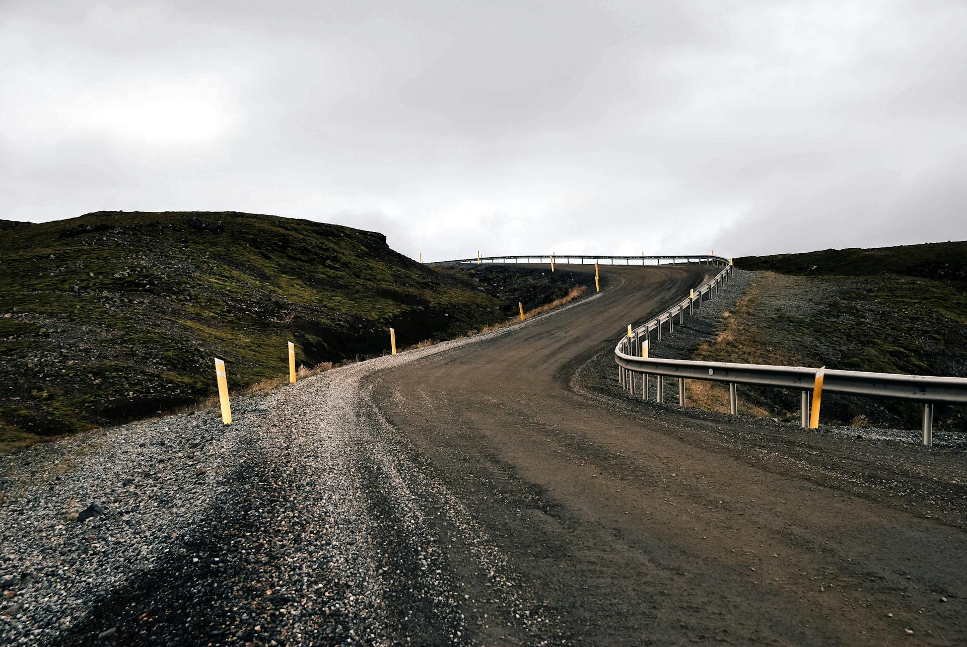 Curved gravel road leading uphill in a rugged rural landscape under cloudy skies.