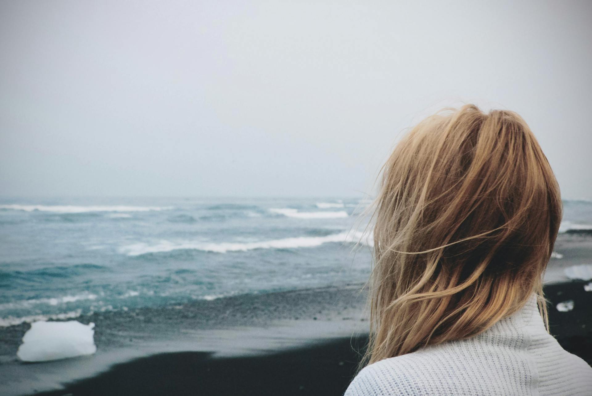 Woman gazing at the ocean on a windy day by a black sand beach with icebergs.