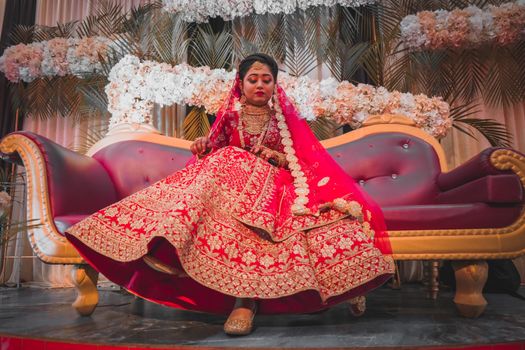 A stunning Indian bride in a red bridal lehenga sitting on a sofa, beautifully adorned with jewelry and makeup.