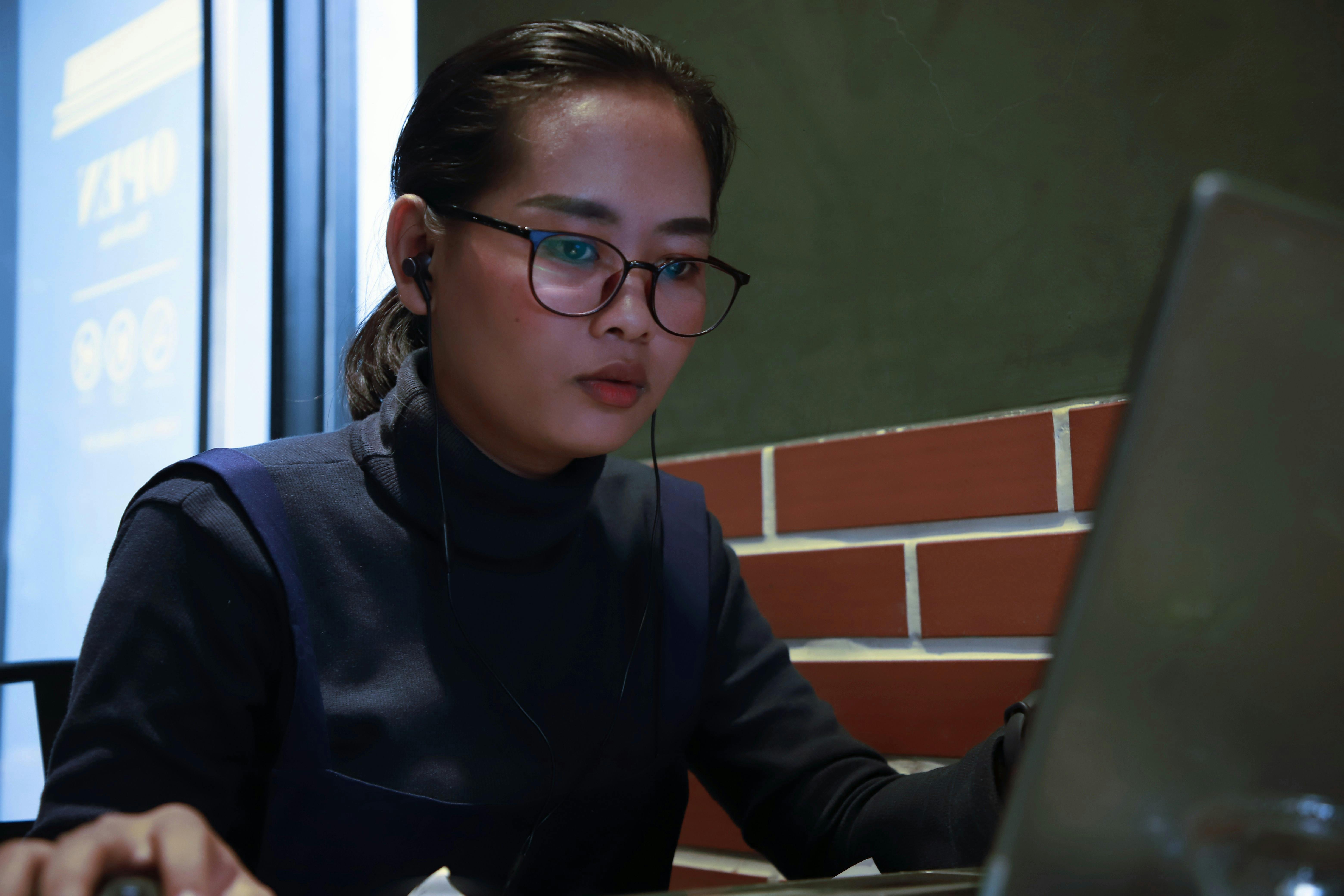 Focused Asian woman in glasses working indoors on a laptop, showcasing concentration.