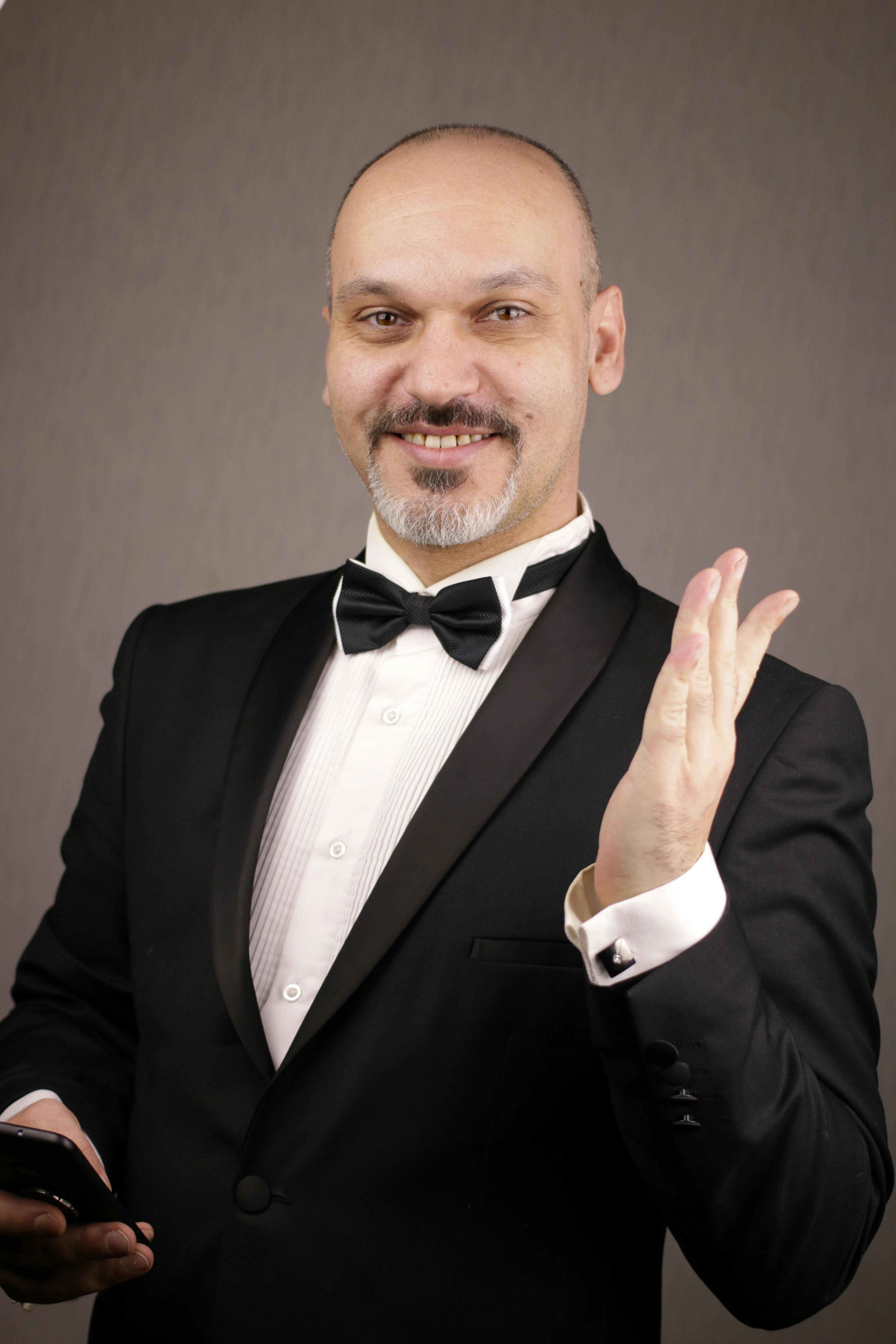 Portrait of a smiling bald man in a black suit holding a cellphone against a studio background.