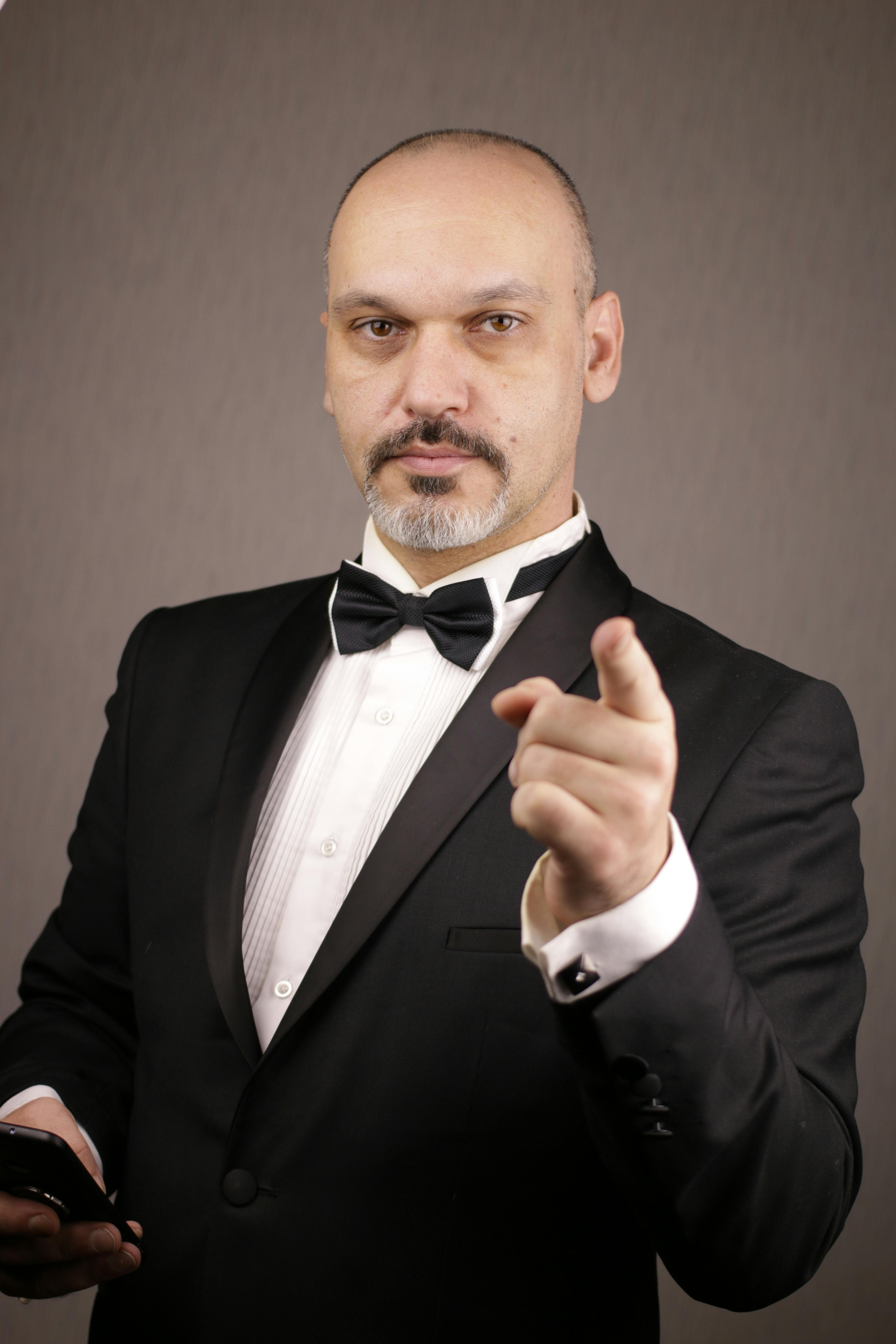 Portrait of a confident bald man in a tuxedo pointing at the camera in a studio setting.