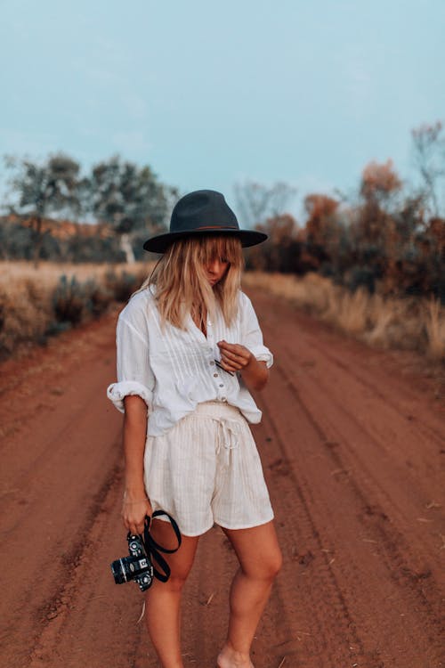 Woman in White Dress Shirt and Brown Hat Standing on Brown Sand Holding a Camera