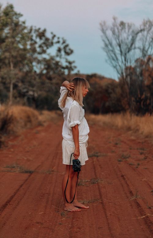 Free A woman in a white shirt holding a camera stands barefoot on a rural dirt road at sunset. Stock Photo