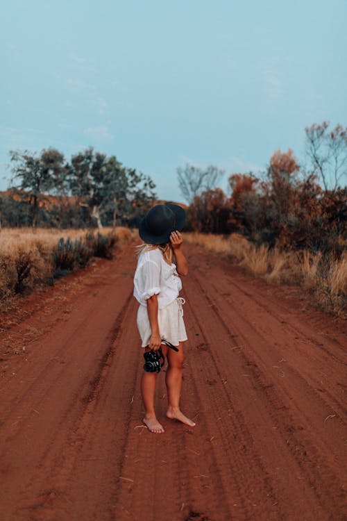 Woman Standing and Holding a Camera in the Middle of an Unpaved Red Sand Road 