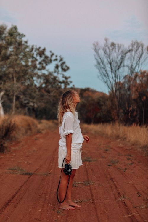 Free Barefoot woman in white dress standing on a dirt road holding a camera during daytime. Stock Photo