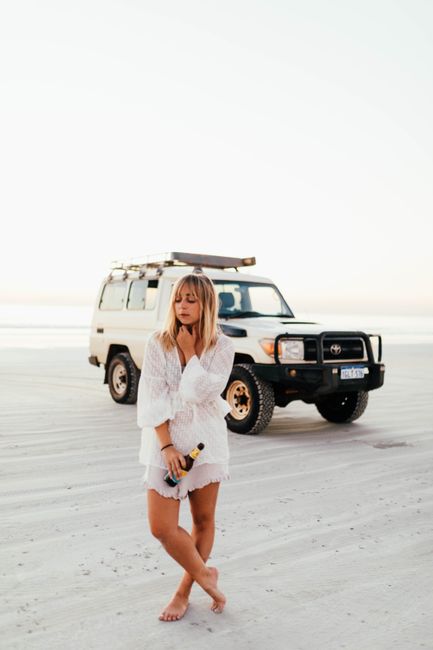 A woman in a white dress stands on a beach holding a bottle, with an SUV in the background.
