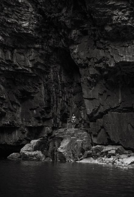 A lone person stands on a rocky cliff above water in a black and white scene.