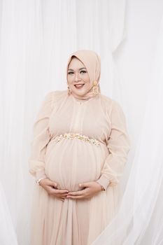 Pregnant woman in a hijab smiling during an elegant studio shoot representing motherhood. - Fotografer Sidoarjo