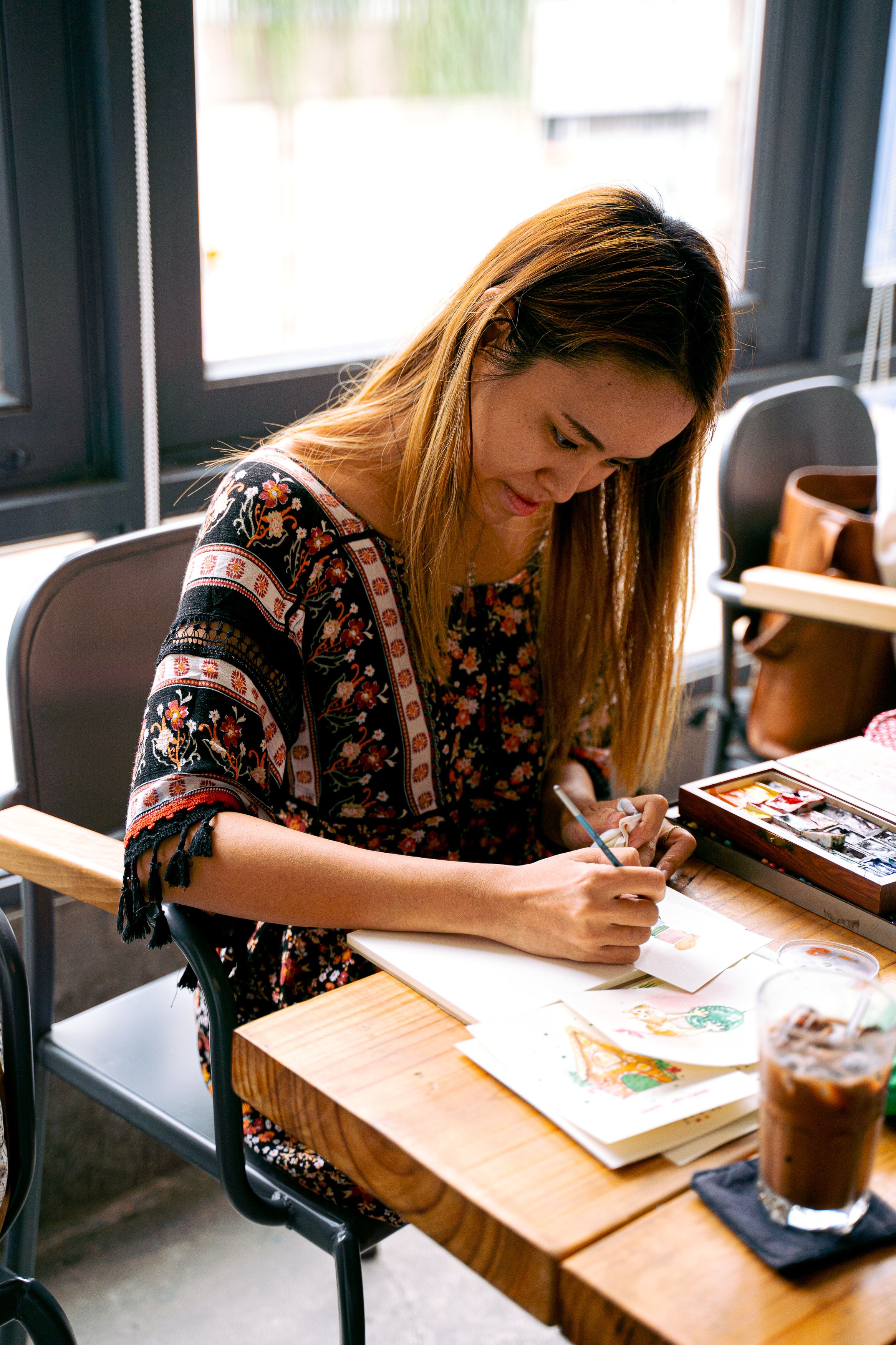 A young woman drawing and painting at a table indoors, showcasing creativity and focus.