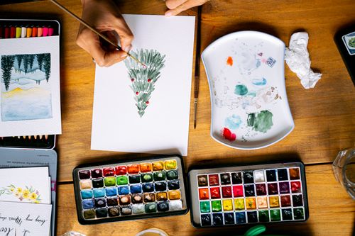Free Artistic workspace featuring a watercolor Christmas tree painting with vibrant paints. Stock Photo