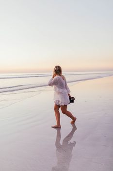 A woman with a camera walks along a serene beach during sunset, reflecting in the wet sand.