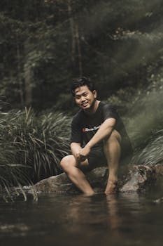 Man sitting by a calm stream in lush greenery, enjoying nature in Kepulauan Riau.